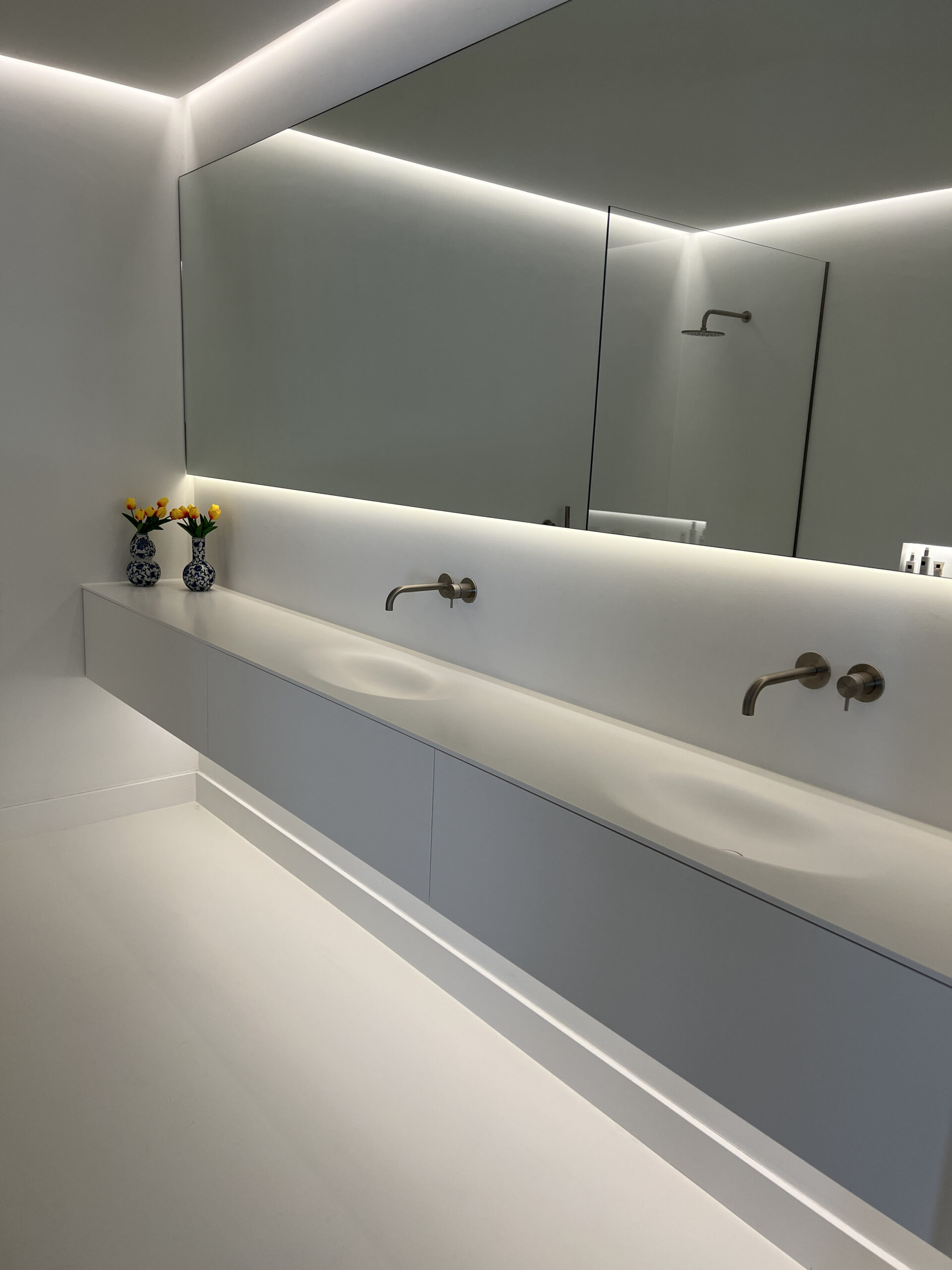 Een witte badkamer met een wit badkamermeubel en een dubbele wastafel waarboven 2 kranen hangen en een grote rechthoekige spiegel met LED verlichting. Op het badkamermeubel staan twee delfstblauwe vaasjes met in ieder een gele bloem